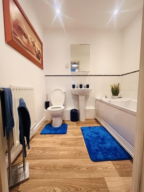 A modern bathroom with a white toilet, sink, and bathtub. There are blue mats on the floor and a framed picture on the wall. - Beautiful 2 - Bed Apartment in Aylesbury (Aylesbury)