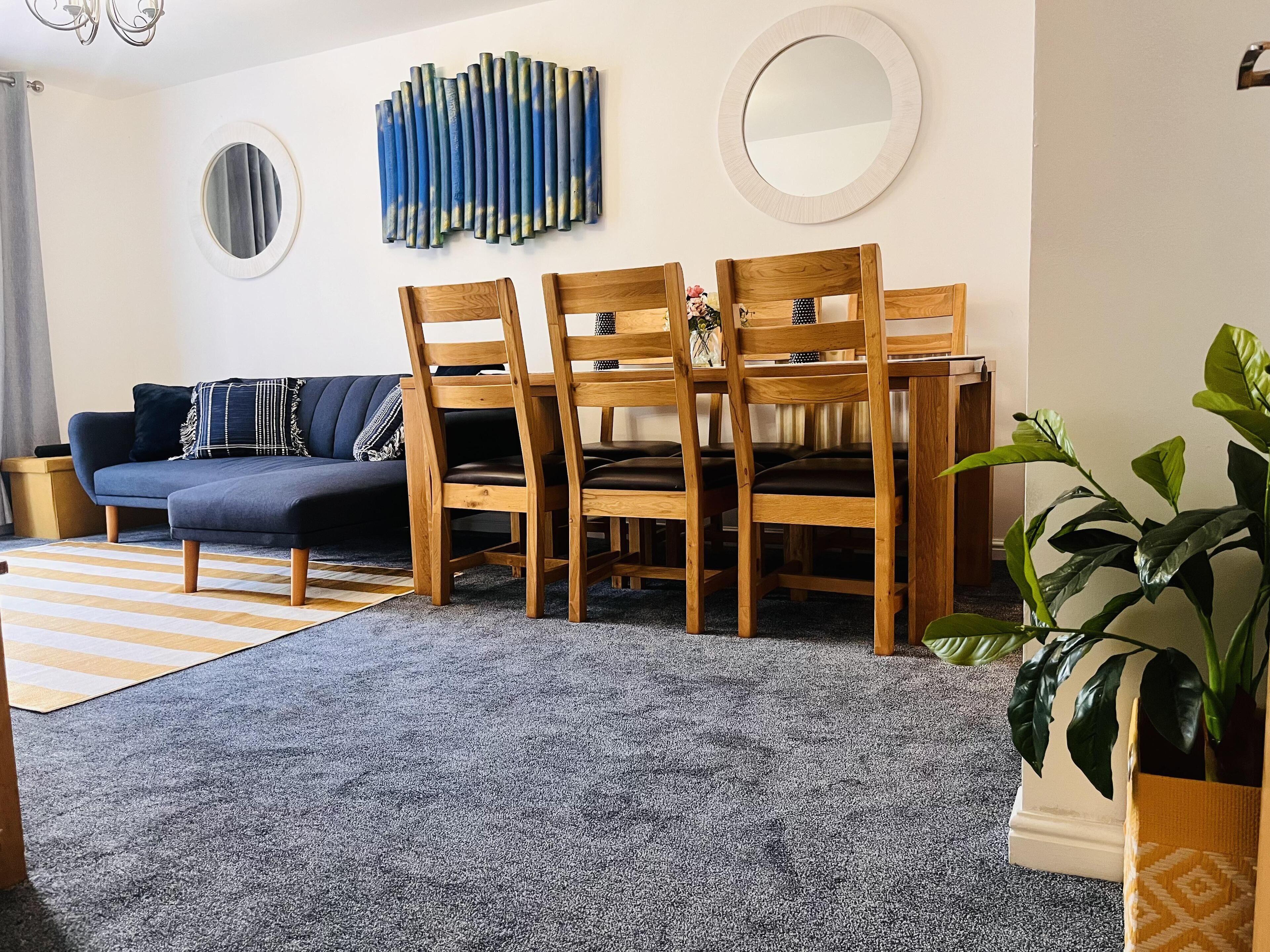 A modern living room with a grey sofa, wooden dining table, and chairs, a grey carpet, and a large plant.