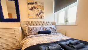 Ein Schlafzimmer mit einer gepolsterten Kopfstütze, floralem Bettzeug, einem großen abstrakten Gemälde, einem Fenster mit Blick und einem Nachttisch mit Lampe.