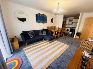 A living room with a dark blue sofa, a striped rug, a chandelier, and a kitchen in the background. - Beautiful 2 - Bed Apartment in Aylesbury (Aylesbury)