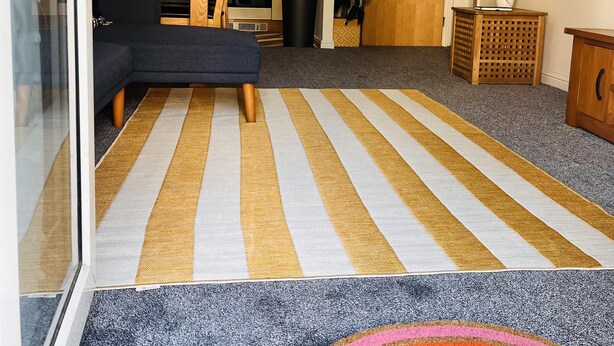 Ein Wohnzimmer mit einer Couch, einem gestreiften Teppich, einem Teppich mit Regenbogenmuster, einer Holzkommode und einer Küche im Hintergrund.