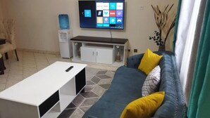 A living room with a sofa, coffee table, TV, and a small cabinet. - Apt 5 - 1BR Apartment with a pool, high-speed internet, close to the airport (Nairobi)