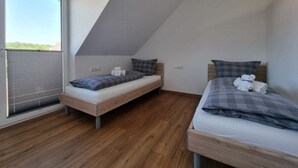 A small, modern bedroom with two single beds, a large window with blinds, and a sloped ceiling.