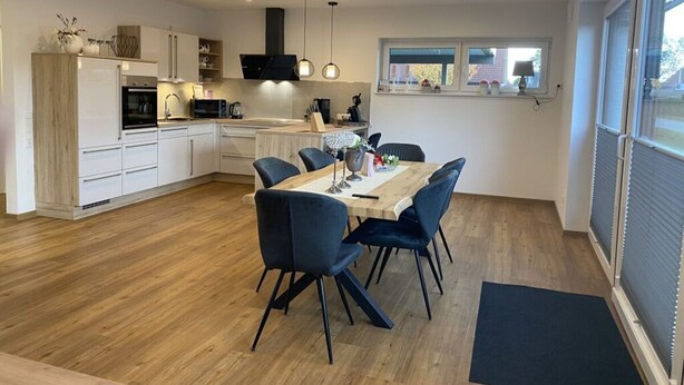 A modern kitchen with white cabinets, a dining area with a wooden table and blue chairs, and a large window providing natural light.
