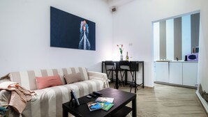 A living room with a striped sofa, a coffee table with books and remote controls, a painting on the wall, and a kitchen area in the background. - Lake Como Love (Como)