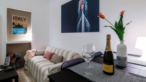 A living room with a sofa, a glass of wine, a bottle of wine, a vase with flowers, a framed picture of Italy, and a painting of a rocket launch. - Lake Como Love (Como)