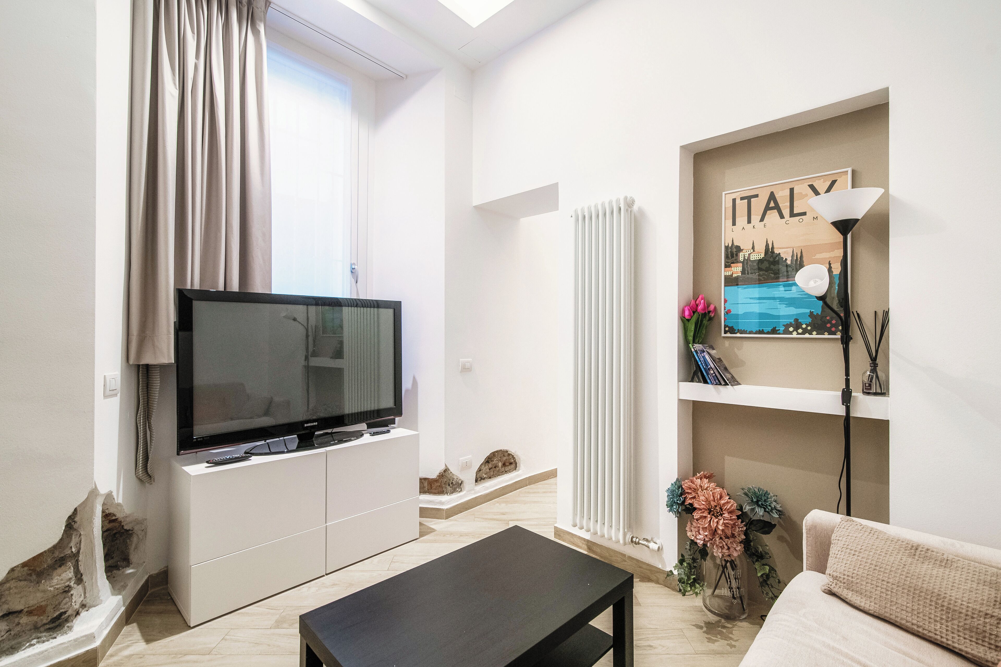 Una sala de estar moderna con un televisor de pantalla plana sobre un mueble blanco, una mesa de centro negra, un sofá color beige y un radiador fijado a la pared.