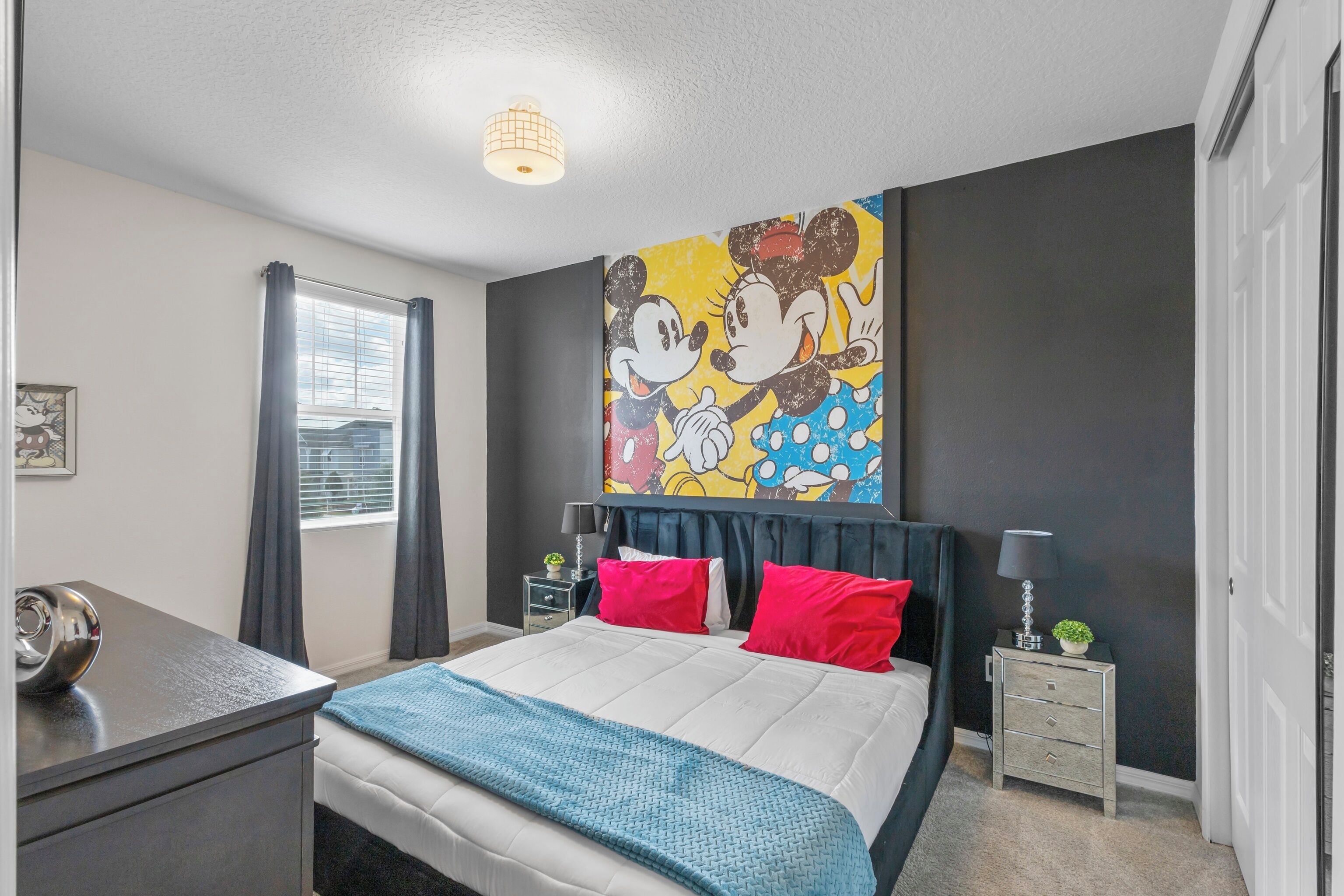 Une chambre avec un lit, un bureau, une table de chevet et une grande fresque murale représentant Mickey et Minnie Mouse.