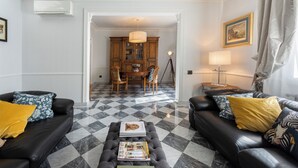 A living room with two black leather sofas, a coffee table with magazines, a patterned rug, a dining area with wooden cabinets, and a chandelier. - Ippopotamo by Mmega (Florence)