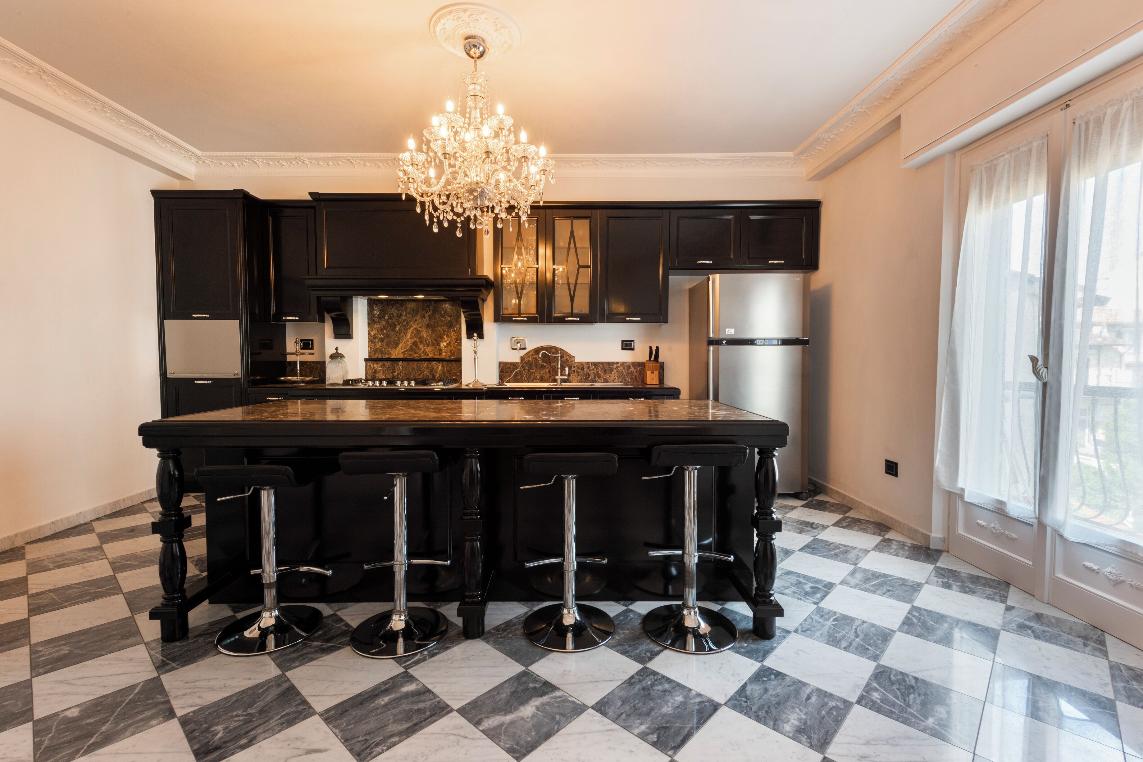 A kitchen with dark cabinets, a marble countertop, a central island with bar stools, a stainless steel refrigerator, and a chandelier.