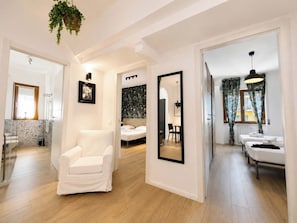 A modern hotel room with a white armchair, a bed with a headboard, a mirror, and a dining area with a table and chairs.