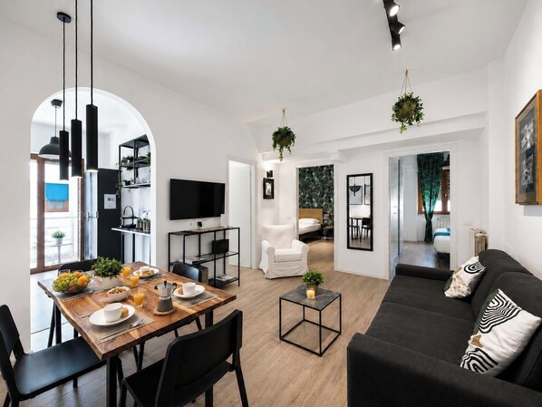 A modern living space with a dining area, a sofa, a TV, and a kitchen visible through an archway.