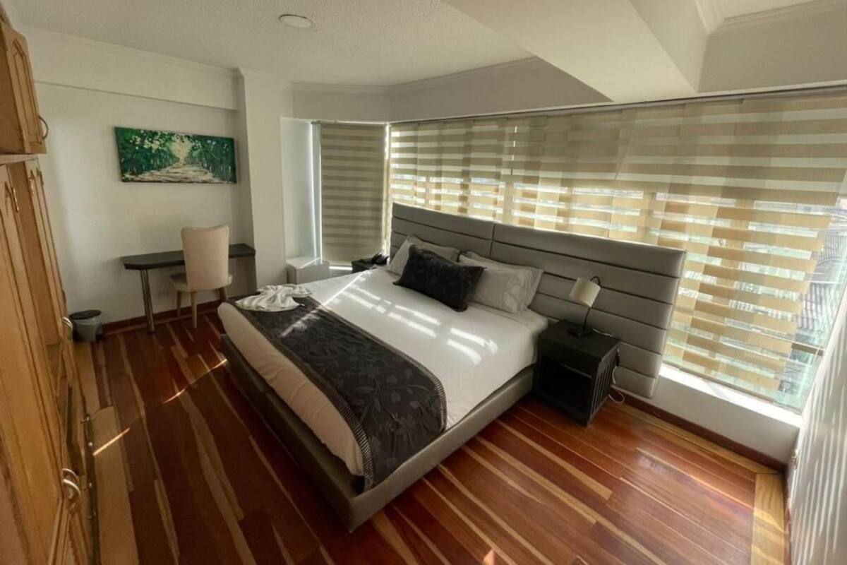 A modern bedroom with a large bed, a bedside table, a chair, and a window with blinds.