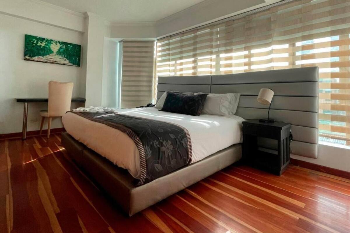 A bedroom with a large bed, a bedside table, a chair, and a window with blinds.