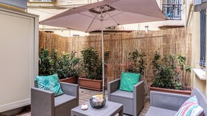 Un patio con mobiliario de mimbre, una mesa y un parasol grande. Se ven plantas en macetas y una valla de bambú al fondo.