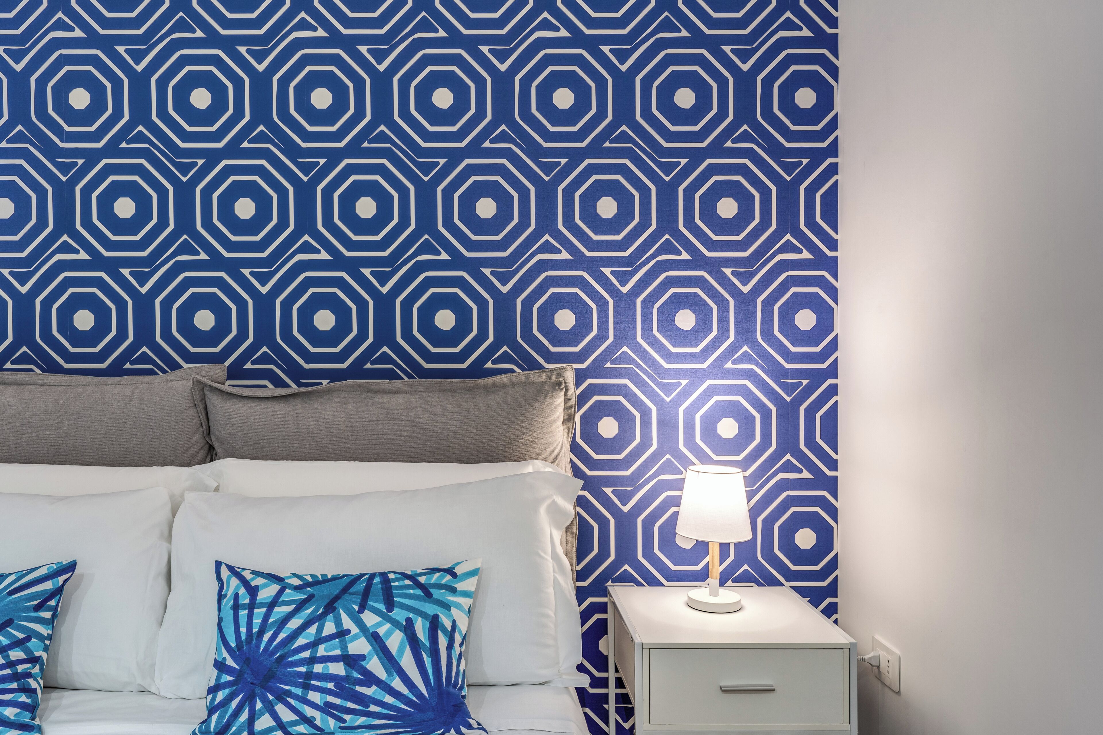 Una cama bien hecha con un papel tapiz de diseño geométrico, una mesita de noche con una lámpara y un enchufe en la pared.