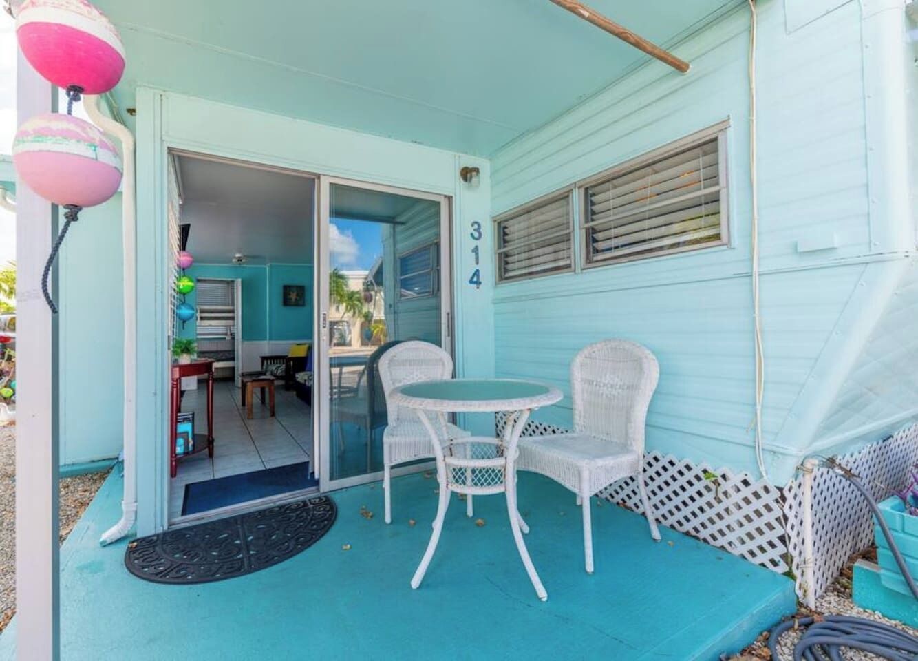 Une maison de couleur turquoise avec le numéro « 314 » peint sur le mur, une table en osier blanche avec deux chaises, et un petit espace de véranda.