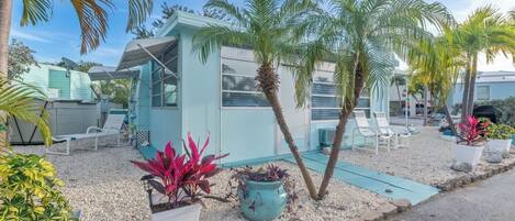A modern, light blue building with a covered outdoor seating area, surrounded by palm trees and potted plants.