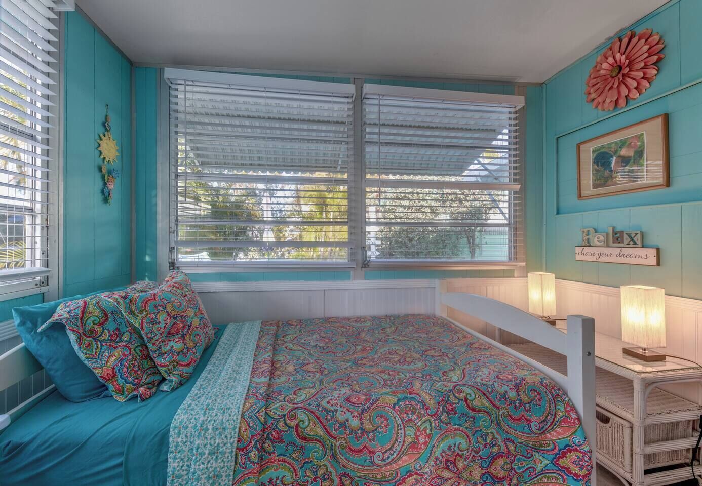A bedroom with a bed, a window with blinds, a wall decoration, and a bedside table with a lamp.