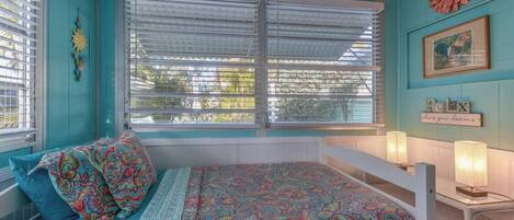 A bedroom with a bed, a window with blinds, a wall decoration, and a bedside table with a lamp.