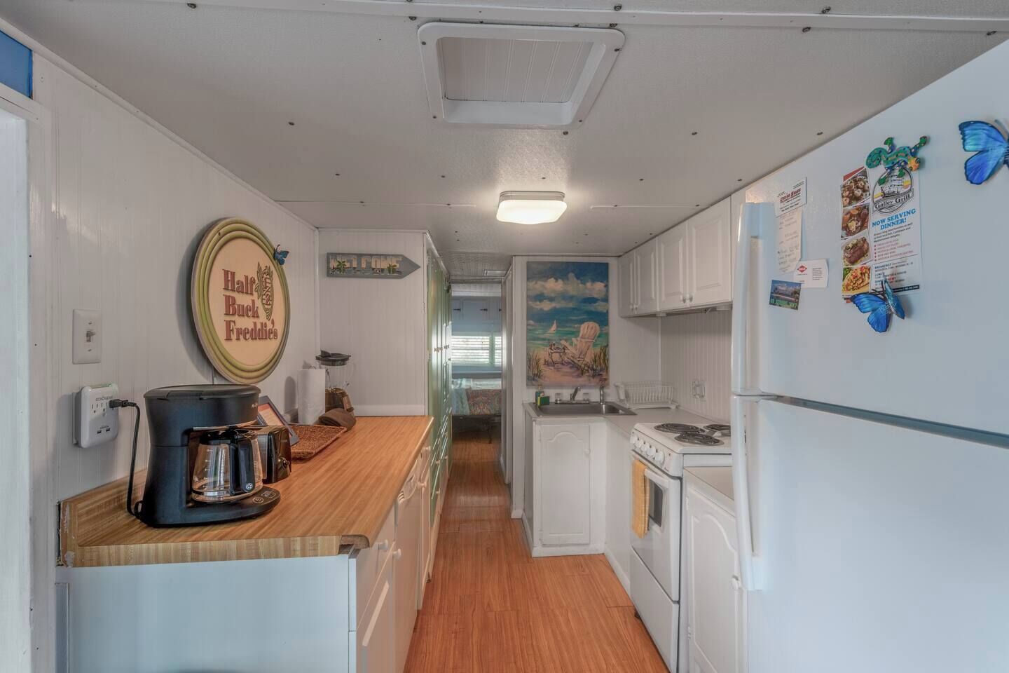 Une petite cuisine avec des meubles blancs, une machine à café et une cuisinière. Une pancarte sur le mur indique : « Hal's Bucky's ».