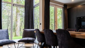 A modern dining area with a wooden table and dark chairs, large windows with curtains, and a kitchen with a black oven. - Elegant Country Chalet in Guelders (Hoenderloo)