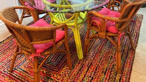 Ein Essbereich mit einem runden Tisch mit Glasplatte, umgeben von Korbstühlen mit rosa Kissen. Ein farbenfroher Teppich bedeckt den Boden, und auf dem Tisch steht eine Vase mit gelben Blumen.