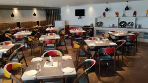 A restaurant with multiple tables and chairs, some with colorful cushions. Tables are set with plates, glasses, and cutlery. - Hotel Southern Grand (Vijayawada)