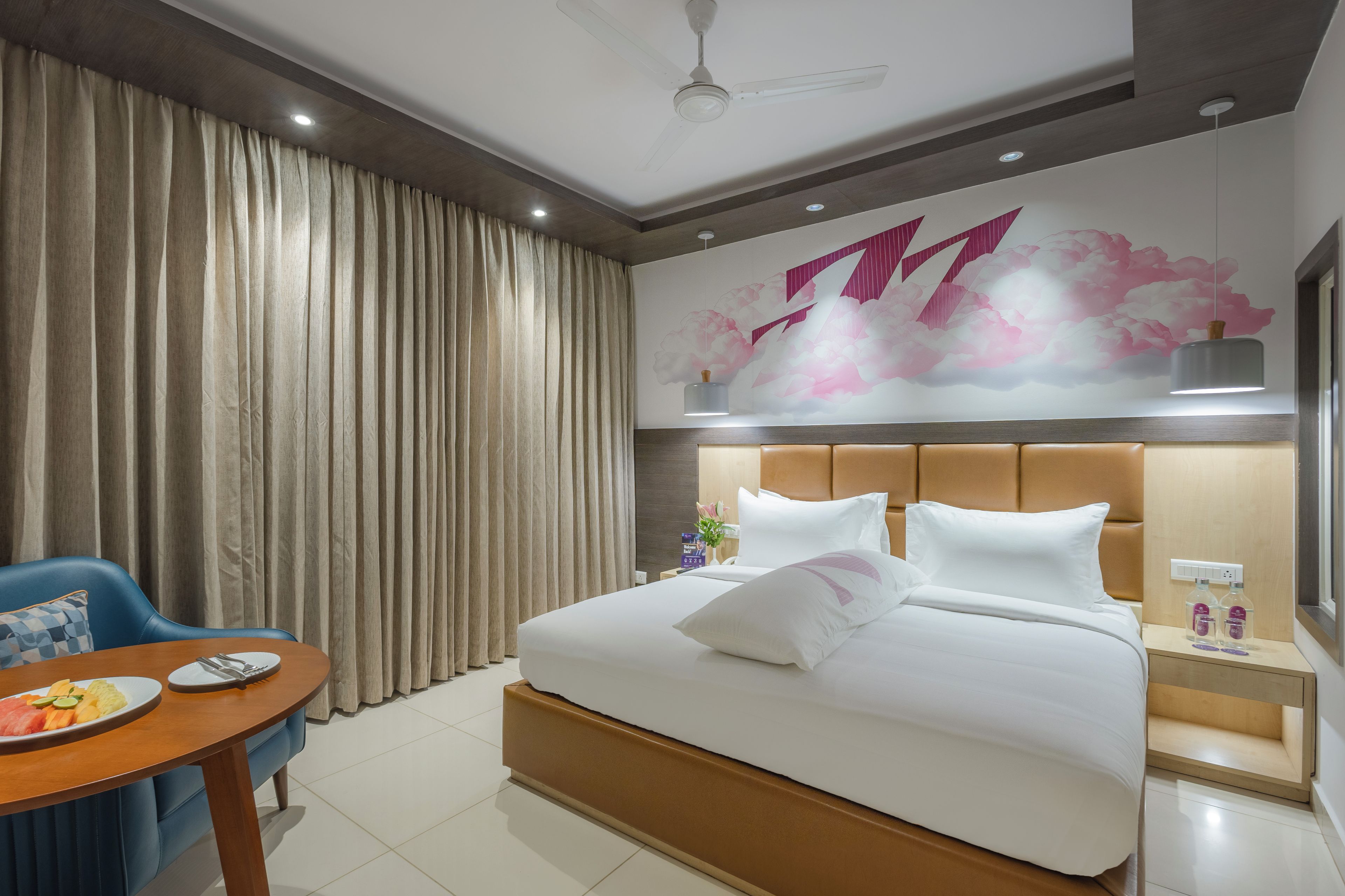 A hotel room with a bed, a desk with a chair, a wall with a floral mural, and a window.