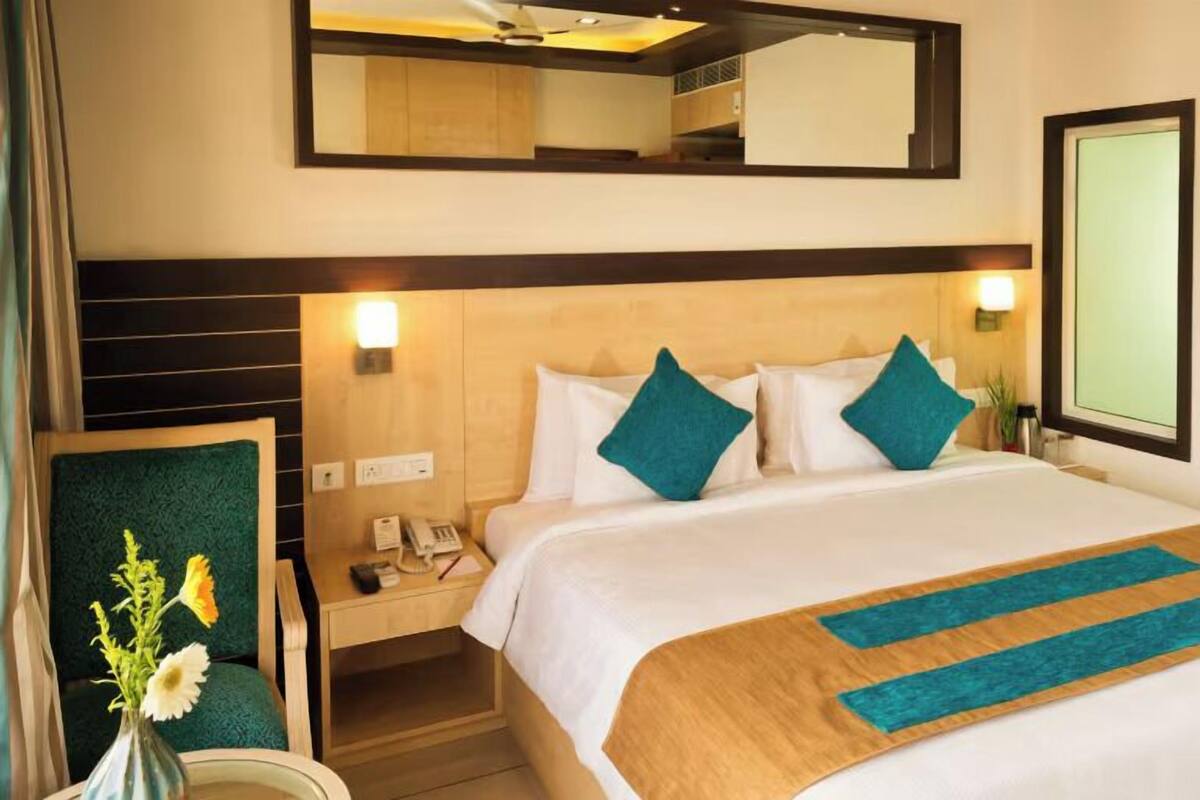 A hotel room with a bed, a nightstand, a mirror, a wall-mounted TV, and a vase with flowers.