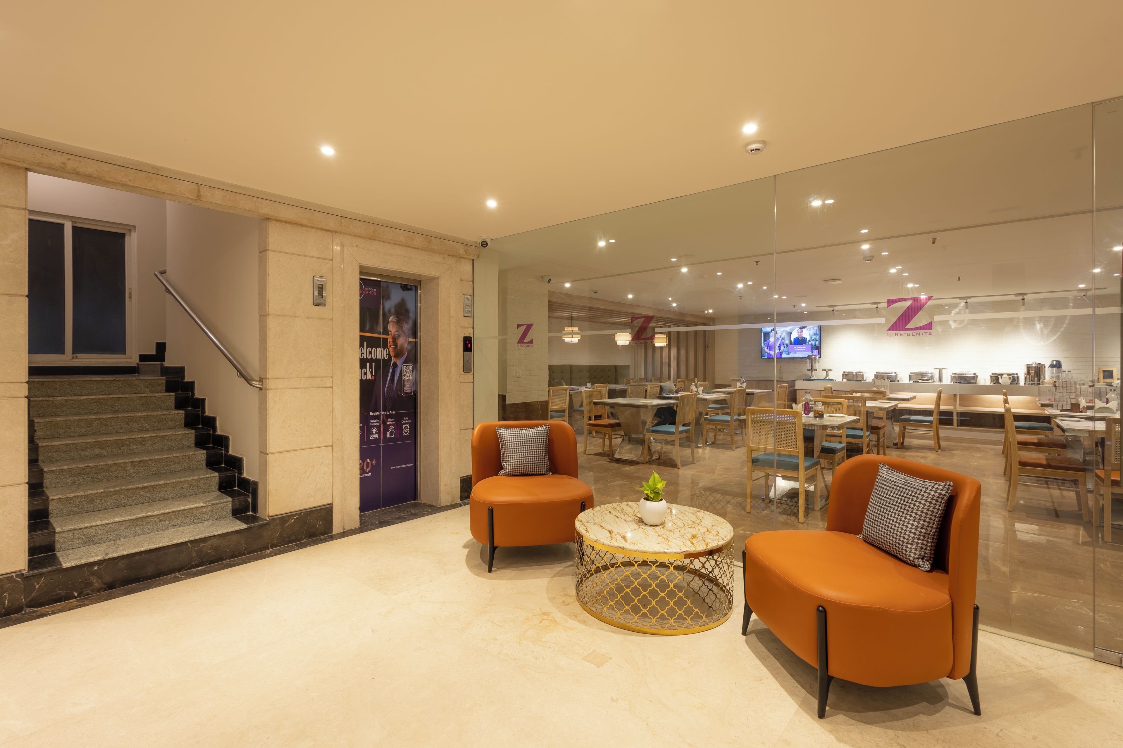 A hotel lobby with a staircase, seating area with orange chairs, a coffee table, and a reception desk with a TV screen.