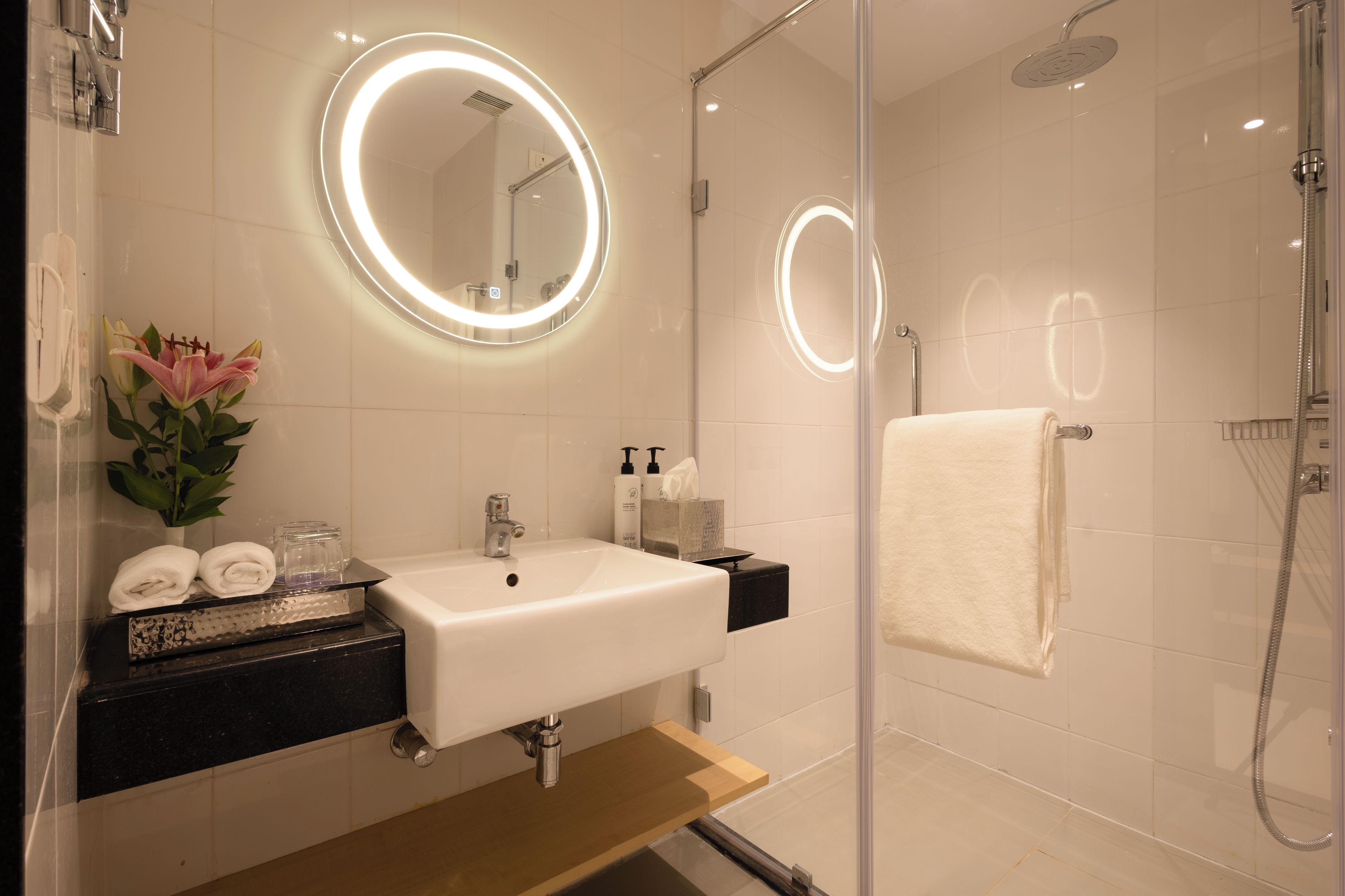 A modern bathroom with a glass-enclosed shower, a white sink, and a mirror with a circular light.