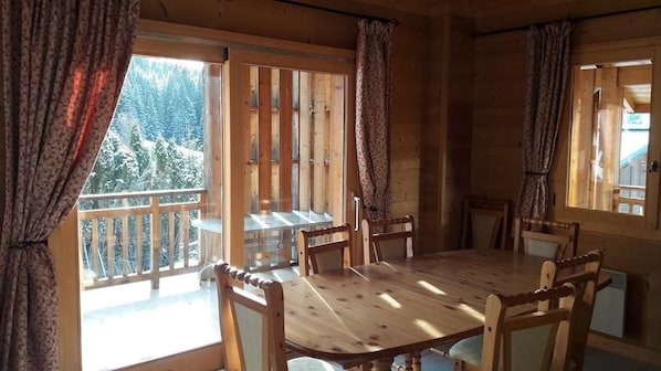 A wooden dining table with six chairs, a balcony with a wooden railing, and a view of snow-covered trees.