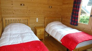 A wooden cabin room with two beds, each with a white duvet and a red blanket. There is a small wooden nightstand between the beds.
