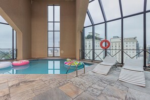 En indendørs swimmingpool med krystalklart blåt vand, stenbelagte gulve, liggestole og udsigt til byens bygninger gennem store vinduer.