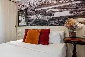 Una cama bien hecha con almohadas blancas y rojas, un cuadro en la pared y un florero con flores secas sobre una mesita de noche.
