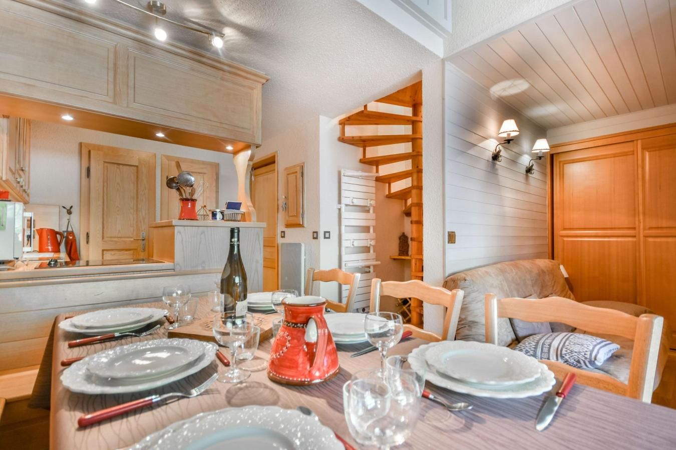 A dining area with a wooden table set for four, surrounded by wooden chairs. A spiral staircase is visible in the background, leading to an upper level.