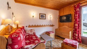 Una habitación acogedora con techo de madera, una cama con colcha roja estampada, un televisor montado en la pared y una ventana con cortinas rojas.