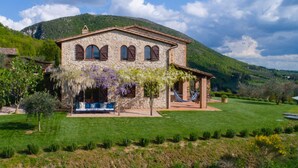 Un bâtiment en pierre doté d’un porche, d’un hamac et d’un banc, situé dans un espace verdoyant avec des montagnes en arrière-plan.