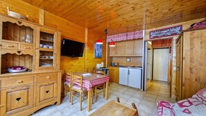 Interior de uma cabana de madeira com mesa de jantar, cadeiras, cozinha equipada com frigorífico e uma televisão na parede.