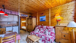Interior acolhedor de uma cabana, com teto de madeira, uma cama beliche, um sofá com manta estampada, mesa de jantar com cadeiras e uma área de cozinha.