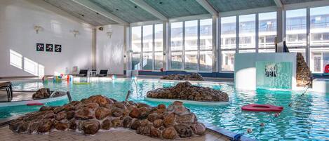 An indoor swimming pool with clear water, rock formations, and a view of the outside through large windows.