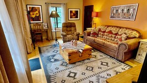 Una sala de estar acogedora con un sofá, un sillón y una mesa de centro con una manta. Hay cuadros enmarcados en las paredes y una lámpara sobre una mesita auxiliar.