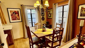 Un comedor con una mesa de madera, sillas y una araña. Hay cuadros en las paredes y una ventana con cortinas.