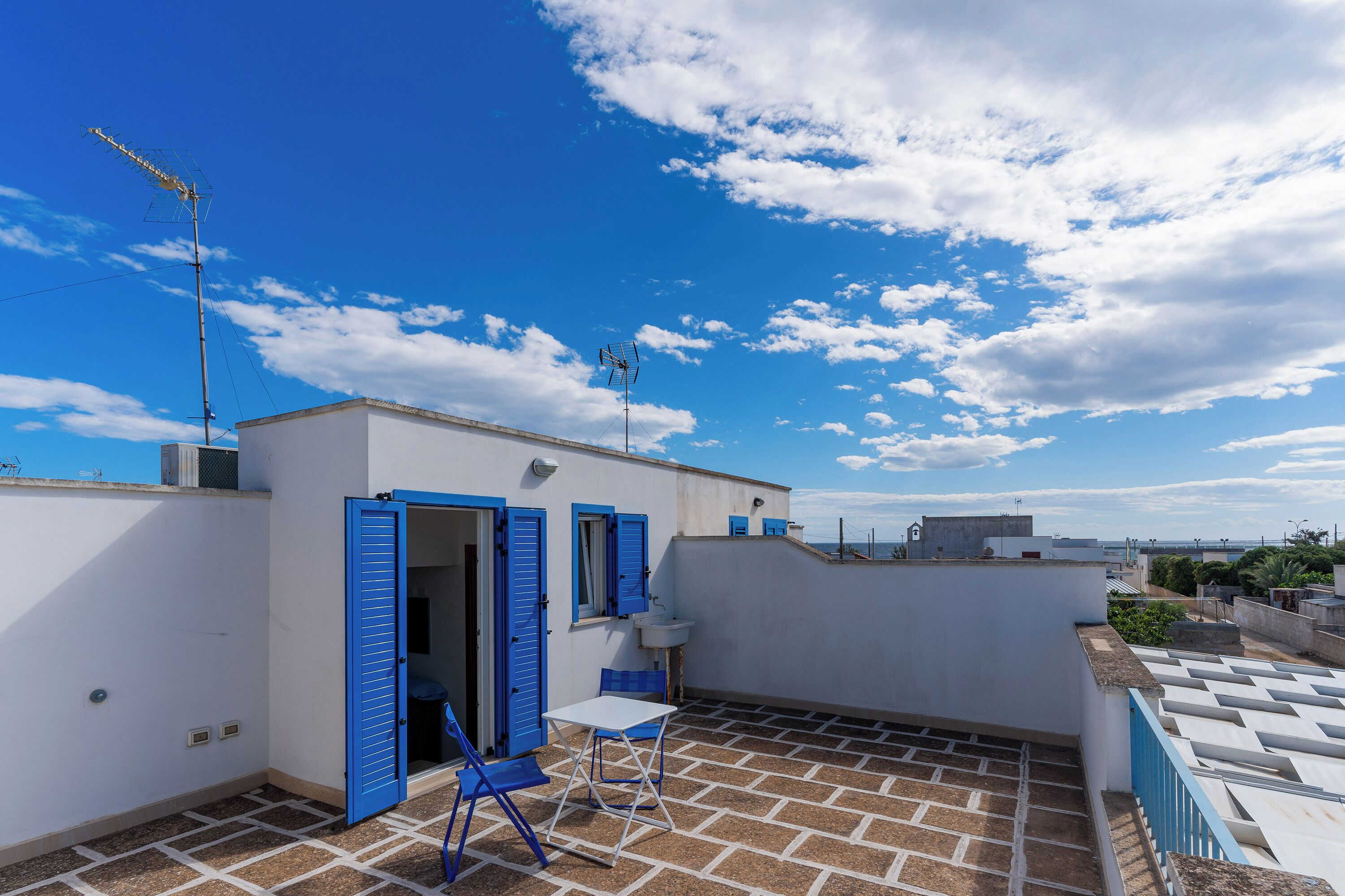 Eine Dachterrasse mit einem weißen Gebäude, blauen Fensterläden, einem Tisch mit Stühlen und Blick zum Himmel.