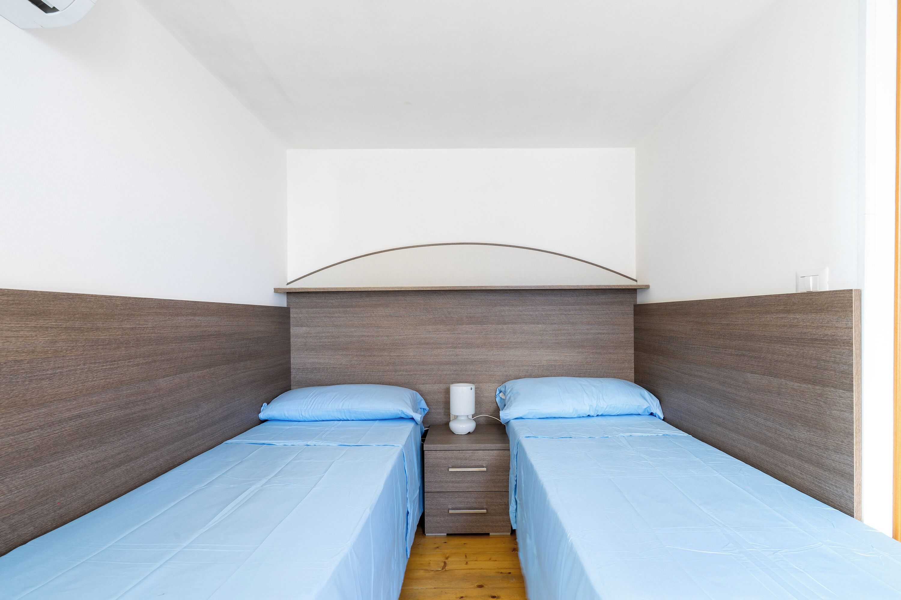 A small, modern bedroom with two single beds, a nightstand, and a wall-mounted lamp.