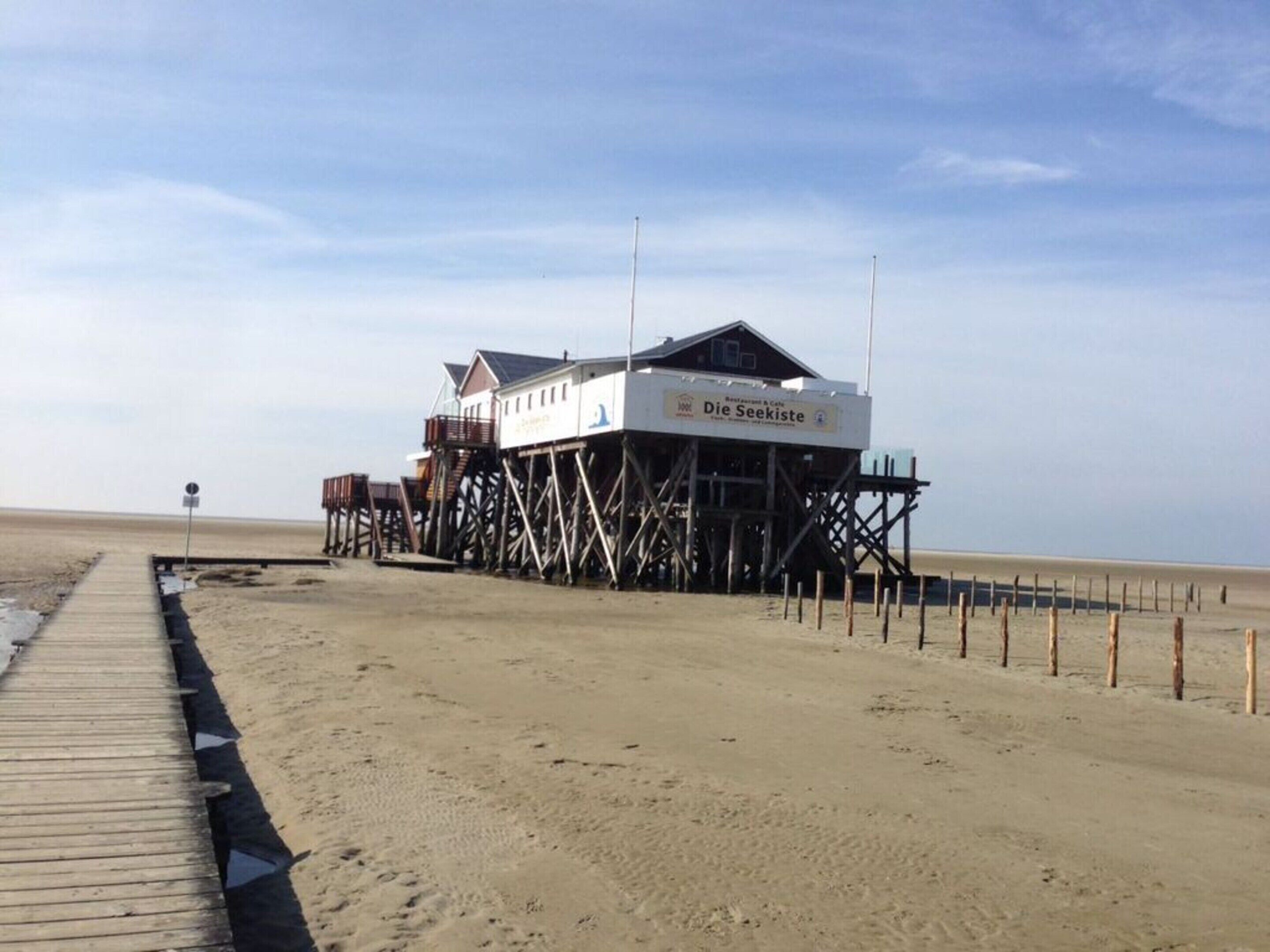 A wooden pier leading to a beach house named 'Die Seekiste' with a restaurant and cafe.