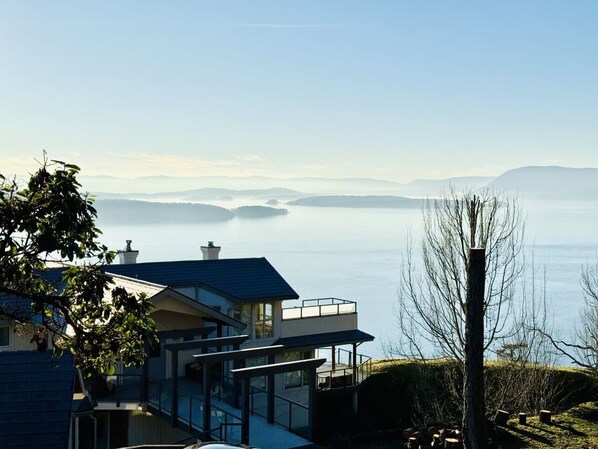 Een modern huis met een glazen gevel, een balkon en uitzicht op een rustig meer met bergen in de verte.