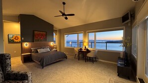 Ein Schlafzimmer mit Bett, Schreibtisch, Stuhl, Deckenventilator und großen Fenstern mit Blick auf das Wasser.
