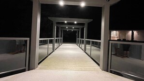 Ein überdachter Holzsteg mit Glasgeländern, der zu einem Gebäude mit sichtbarer Küchenzeile führt.